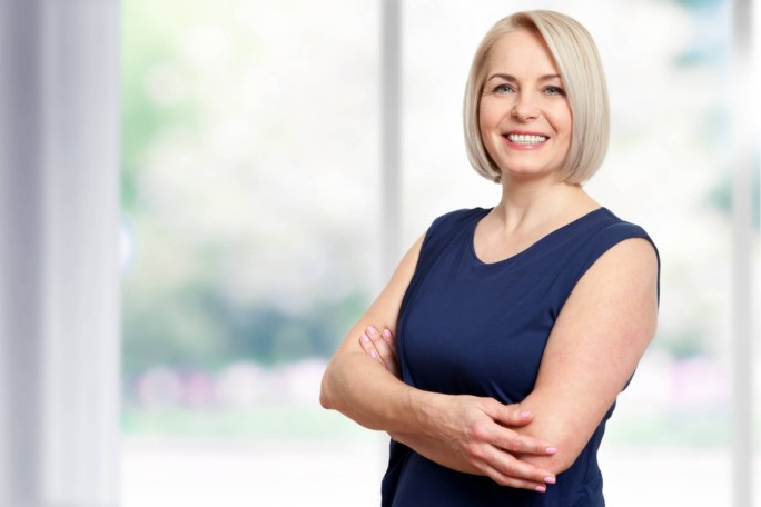Eine Frau in einem ärmellosen, marineblauen Top steht mit verschränkten Armen in einem Raum. Im Hintergrund ist eine leicht verschwommene Ansicht von Grün und großen Fenstern zu sehen, was auf eine helle und luftige Atmosphäre hindeutet. Das Bild vermittelt eine lässige und selbstbewusste Stimmung.
