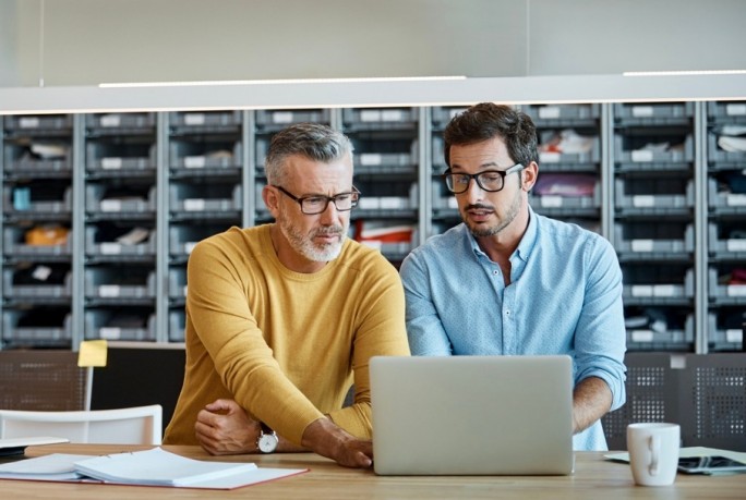 Businessmen using laptop together at desk. Executives are working at textile factory. Male professionals are wearing smart casuals in factory.
