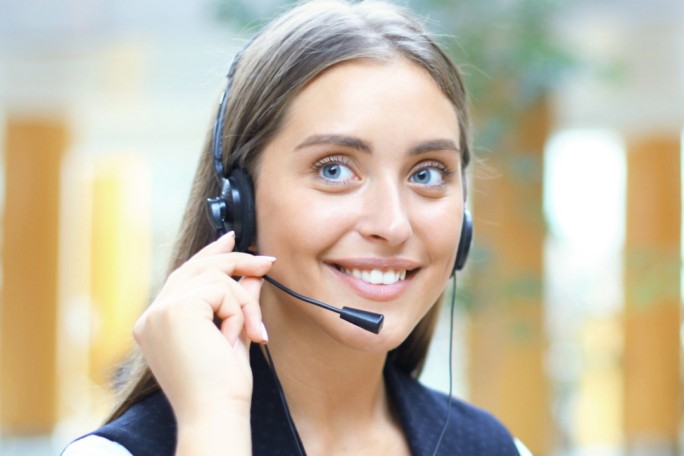 A person is seen wearing black headphones, holding one side with their hand. The setting appears to be indoors with blurred natural light and wooden structures in the background. The image conveys a casual and focused mood.