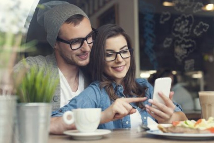 A cozy cafe scene featuring two individuals seated at a table. The focus is on a smartphone being used, surrounded by coffee cups, a plate of food, and a potted plant. The background includes a chalkboard-style wall with visible doodles and text, enhancing the casual and relaxed atmosphere.