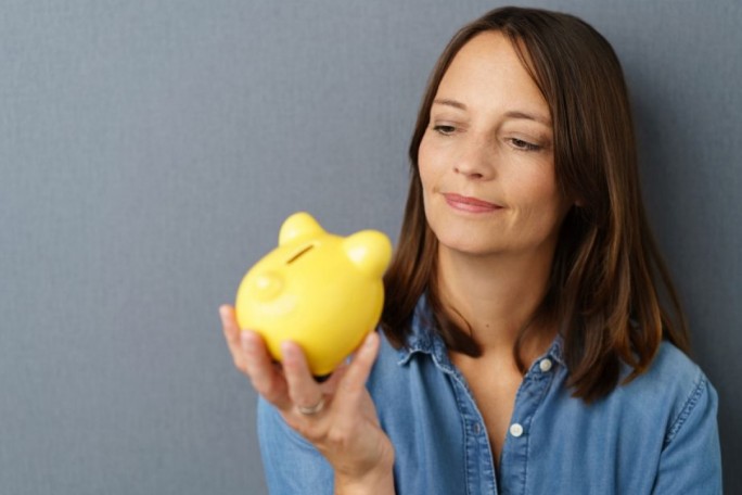 A person holds a bright yellow piggy bank in their hand, symbolizing savings or financial planning. The setting features a neutral gray background and casual attire, creating a relaxed and approachable mood. The piggy bank has a playful design, resembling a cartoonish animal.