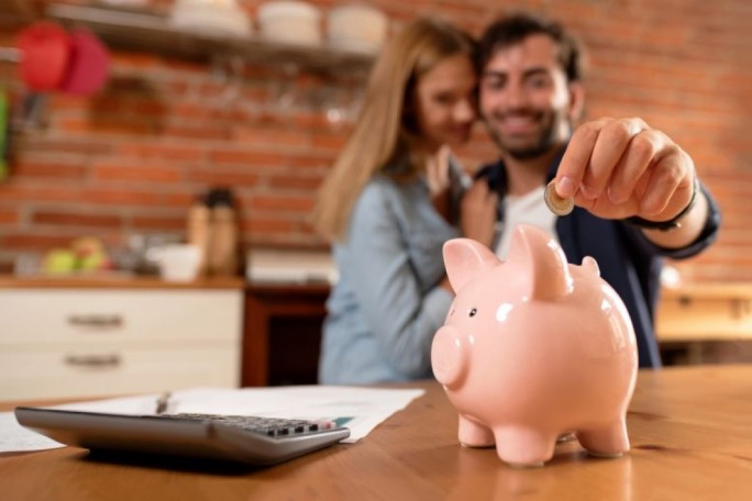 A hand places a coin into a pink piggy bank on a wooden table in a cozy kitchen setting. A calculator and papers are visible nearby, suggesting financial planning or budgeting. The background features a brick wall and kitchen utensils, adding a warm and casual atmosphere.