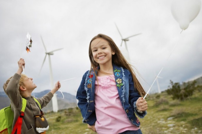 Two children are seen outdoors in a grassy area with wind turbines in the background. One child holds a white balloon, while the other raises a small object resembling a toy. The setting is open and natural, with cloudy skies and a focus on renewable energy visuals.