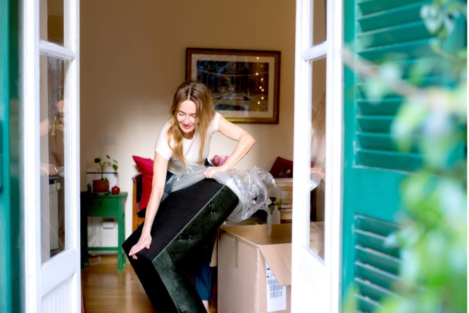 Eine Frau packt in einem gemütlichen Wohnzimmer ein dunkelgrünes Möbelstück aus, vermutlich einen Sessel oder ein Sofa. An der Wand hängen ein gerahmtes Bild, ein kleiner Tisch mit Pflanzen und Kartons, was auf einen Umzug oder eine Lieferung hindeutet. Die Szene wird durch offene Türen mit grünen Fensterläden beobachtet, was dem Raum eine frische und einladende Atmosphäre verleiht.