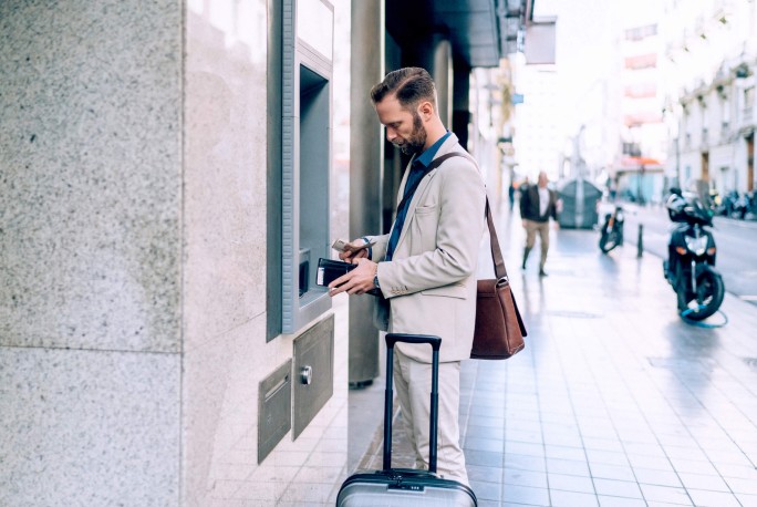 Ein Mann in einem hellen Anzug benutzt einen Geldautomaten auf offener Straße. Er hält Geldbörse und Karte in der Hand und steht neben einem Rollkoffer. Die städtische Umgebung mit gefliestem Gehweg, geparkten Motorrädern und Gebäuden im Hintergrund deutet auf einen Reise- oder Geschäftskontext hin.