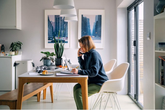 Eine Frau sitzt an einem Holztisch in ihrem modernen Homeoffice und telefoniert, während sie in einem Notizbuch schreibt. Der Raum ist hell und lichtdurchflutet, mit Zimmerpflanzen und einem gerahmten Stadtbild an der Wand. Laptop, Schreibwaren und eine Tasse Kaffee bilden den Arbeitsplatz und schaffen eine produktive und gemütliche Atmosphäre.
